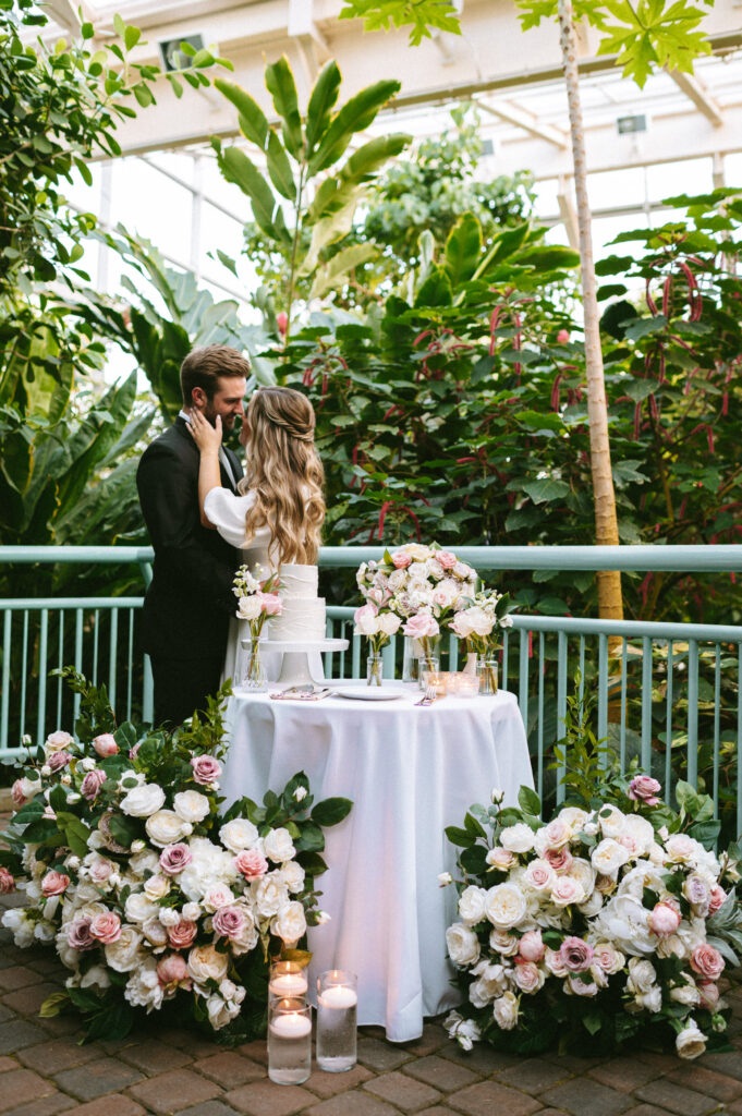 Bride and groom kissing next to wedding cake table surrounded by florals