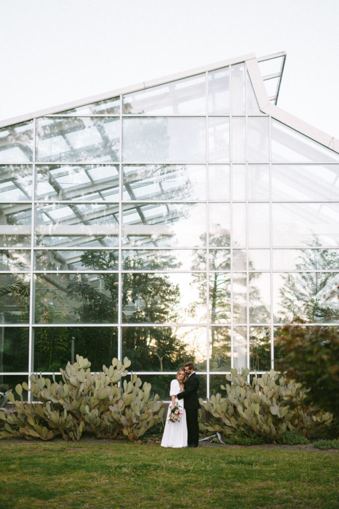 Wide angle view of bride and groom hugging in front of greenhouse building