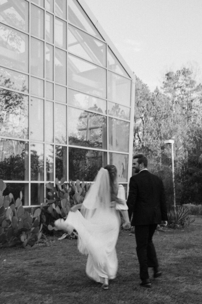 Bride and groom frolicking hand in hand in front of greenhouse