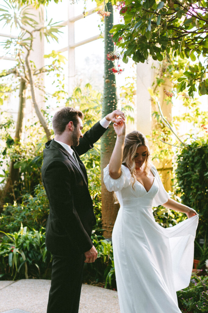 Groom holding bride's hand as she twirls her dress, surrounded by greenery