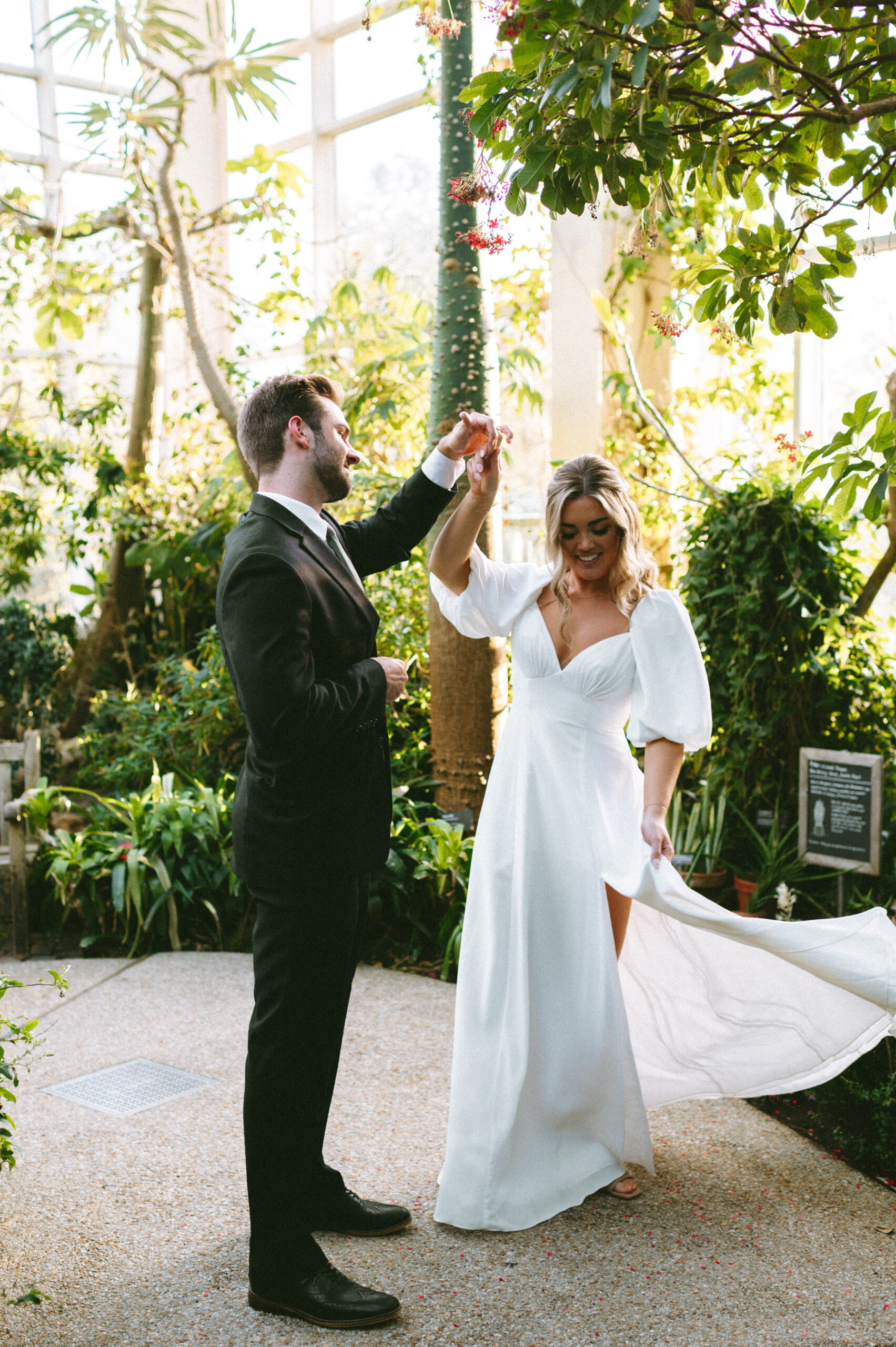 Groom holding bride's hand as she twirls her dress, surrounded by greenery